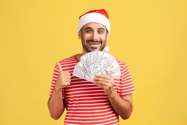 Fröhlicher, zufriedener bärtiger Mann in rotem T-Shirt und Weihnachtsmann-Hut, der Daumen nach oben zeigt, Fan von Dollar hält, mit zahnigem Lächeln in die Kamera schaut, Glück. Studioaufnahme im Innenbereich isoliert auf gelbem Hintergrund