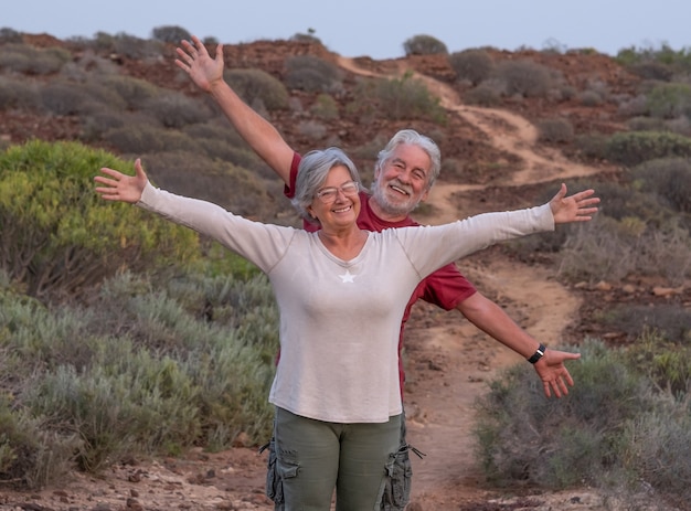 Fröhliches älteres Ehepaar mit ausgestreckten Armen, das einen Ausflug im Freien auf einem Bergwanderweg genießt. Senior entspannte Menschen bei Sonnenuntergang Licht