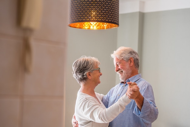 Fröhliches älteres Paar, das tanzt und lächelt und sich ansieht. Älteres glückliches Paar, das durch Tanzen im Wohnzimmer feiert. Romantisches altes Ehepaar, das Händchen hält und zu Hause zusammen tanzt