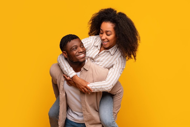 Fröhliches afrikanisch-amerikanisches Paar, das mit Lachen auf gelben Hintergrund auf dem Rücken sitzt