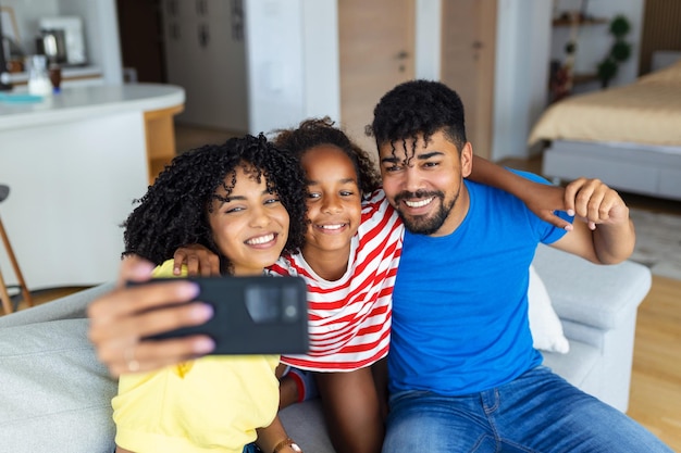 Fröhliches afrikanisches Elternpaar und lachende Tochter, die auf der Couch ruhen und ein Smartphone halten, das ein Selfie auf dem Handy macht und einen Videoanruf macht, der auf den Bildschirm des Mobiltelefons schaut und kichert. Familienfreizeit