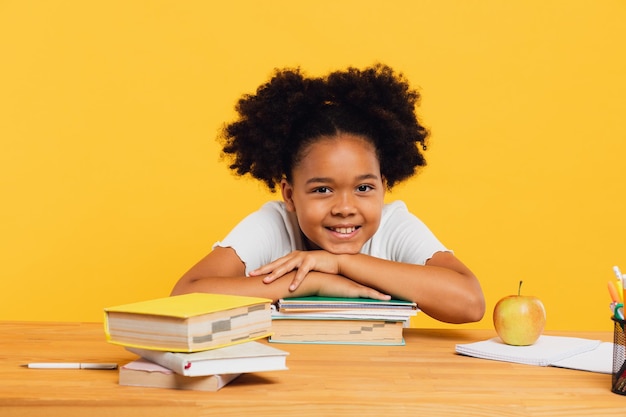 Fröhliches afroamerikanisches Schulmädchen, das am Schreibtisch sitzt und sich auf Bücher stützt Zurück zum Schulkonzept