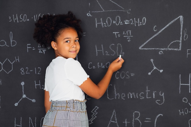 Fröhliches afroamerikanisches Schulmädchen, das Probleme in der Nähe der Tafel in der Schule löst, zurück zum Schulkonzept