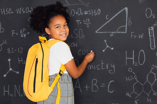 Fröhliches afroamerikanisches Schulmädchen, das Probleme in der Nähe der Tafel in der Schule löst, zurück zum Schulkonzept