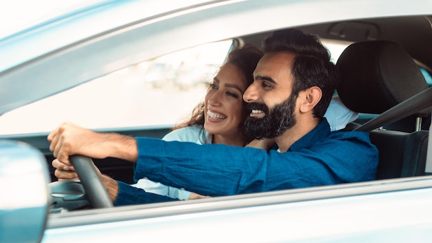 Fröhliches arabisches Paar, das mit geöffnetem Fenster sitzt und sein neues Auto fährt und Straßen erkundet