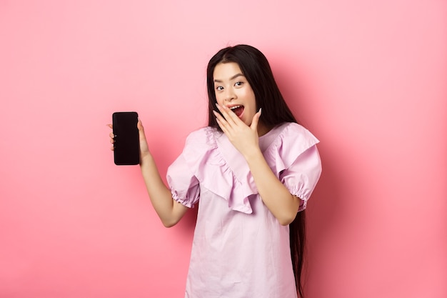 Fröhliches asiatisches jugendlich Mädchen, das leeren Telefonbildschirm zeigt, lacht und Mund mit Hand bedeckt, im Kleid gegen rosa Hintergrund stehend.