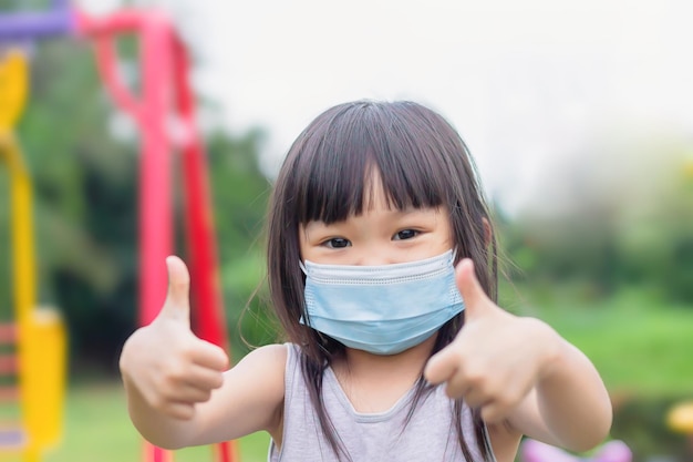 Foto fröhliches asiatisches kindermädchen, das lächelt und eine gesichtsmaske trägt. sie zeigt den daumen nach oben