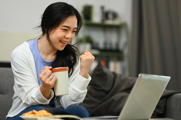 Fröhliches asiatisches Mädchen, das auf den Laptop-Bildschirm schaut und eine Ja-Geste macht, indem es online tolle Neuigkeiten erhält und den Erfolg feiert