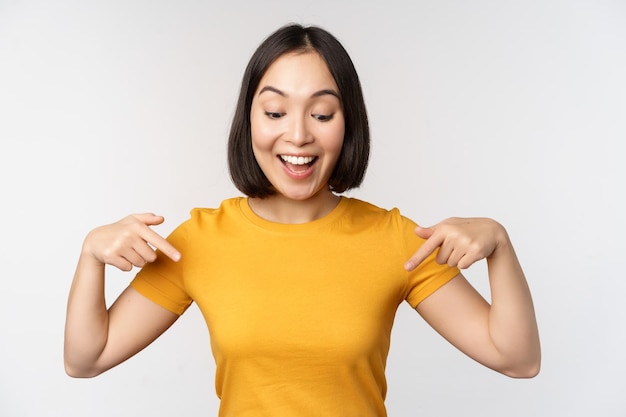 Fröhliches asiatisches Mädchen, das mit dem Finger nach unten zeigt und auf Ankündigungsbanner oder Werbung schaut, die in gelbem T-Shirt auf weißem Hintergrund stehen