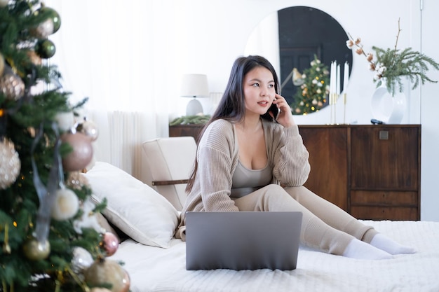 Fröhliches asiatisches Mädchen in gemütlicher Kleidung, das Freunde über das Handy anruft, das zu Hause auf dem Bett sitzt