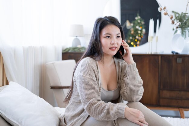 Fröhliches asiatisches Mädchen in gemütlicher Kleidung, das Freunde über das Handy anruft, das zu Hause auf dem Bett sitzt