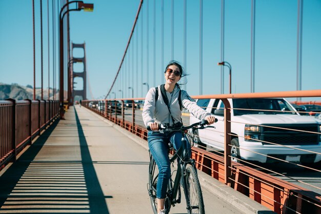 Fröhliches asiatisches Mädchen lacht in die Kamera und hat Spaß beim Fahrradfahren im Sommer auf der Golden Gate Bridge mit vorbeifahrenden Autos in San Francisco USA