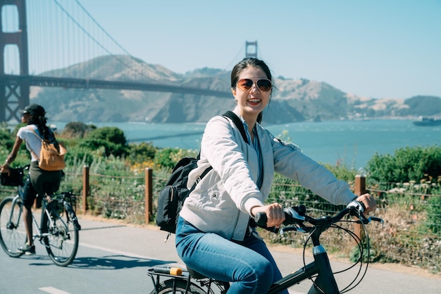 Fröhliches asiatisches Mädchen lächelt auf dem Fahrrad in die Kamera, während es an einem klaren Tag mit Golden Gate Bridge im Hintergrund in San Francisco, Kalifornien, USA, Urlaub macht