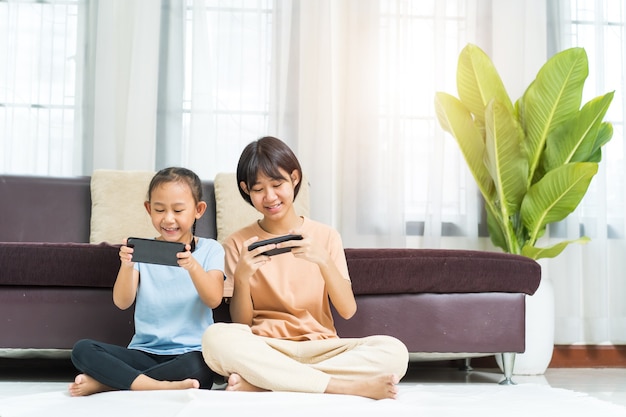 Fröhliches asiatisches Mädchen mit Schwester spielt zu Hause zusammen am Smartphone