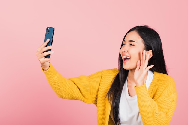 Fröhliches asiatisches Porträt, schöne, süße junge Frau, die aufgeregt lächelt und Selfie-Foto macht, Videoanruf auf Smartphone-Studioaufnahme isoliert auf rosafarbenem Hintergrund, weibliches Handy hebt die Hand und sagt Hallo