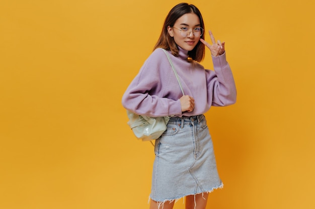 Fröhliches asiatisches Teenager-Mädchen in stylischem Jeansrock und lila Pullover zeigt V-Seufzer und hält Minz-Rucksack an orangefarbener isolierter Wand