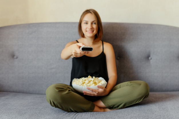 Fröhliches attraktives Mädchen mit Popcorn, das auf Sofa sitzt, fernsieht, Kanäle mit einer Fernbedienung wechselt und Popcorn isst. Zuhause.