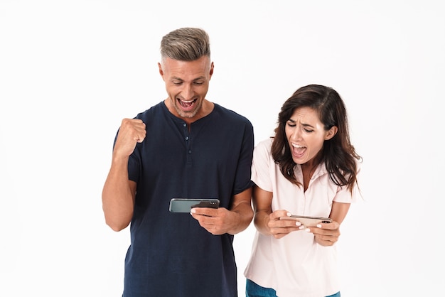 Fröhliches attraktives Paar in lässigem Outfit, das isoliert über weißer Wand steht und Spiele auf Mobiltelefonen spielt