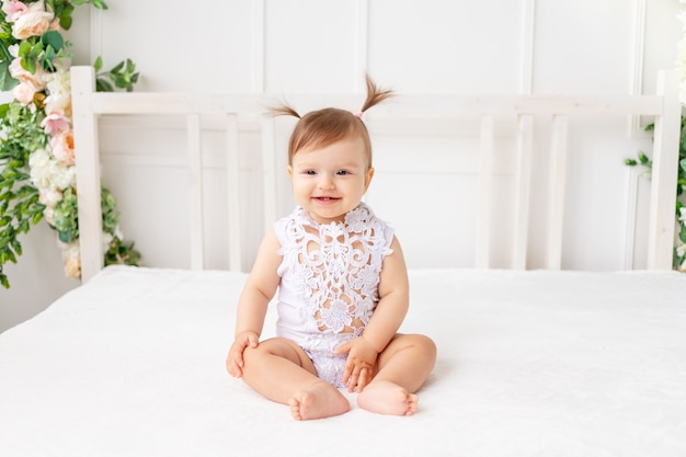 Fröhliches Baby, das in einem hellen schönen Raum auf einem weißen Bett in einem Spitzenbodysuit sitzt und lächelt