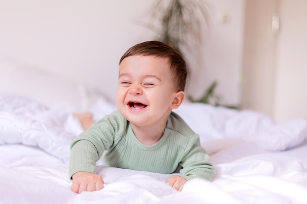 Fröhliches Baby in einem grünen Baumwollbody liegt auf einem weißen Bett auf dem Bauch Hochwertiges Foto