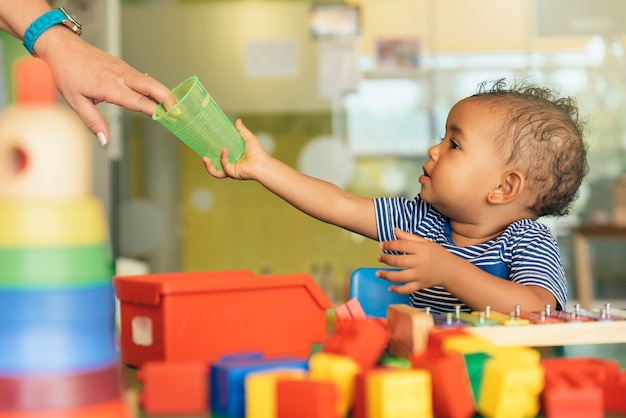 Fröhliches Baby trinkt Wasser und spielt mit Spielzeugblöcken im Kindergarten.