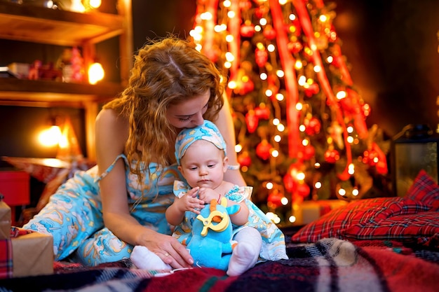 Fröhliches Baby und Mutter sitzen neben dem Weihnachtsbaum im Wohnzimmer Mama küsst kleine Tochter