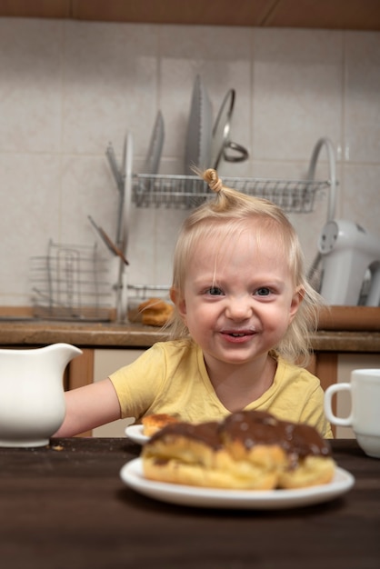 Fröhliches blondes Kind in der Küche frühstückt und betrachtet die Kuchen. Kleines Mädchen, das Tee mit Süßigkeiten trinkt.