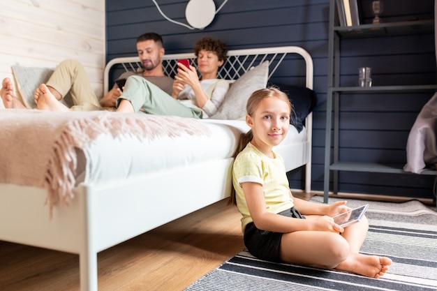 Fröhliches blondes kleines Mädchen mit digitalem Tablet, das auf dem Boden am Bett sitzt, wo sich ihre Eltern entspannen und SMS schreiben oder in ihren Smartphones scrollen