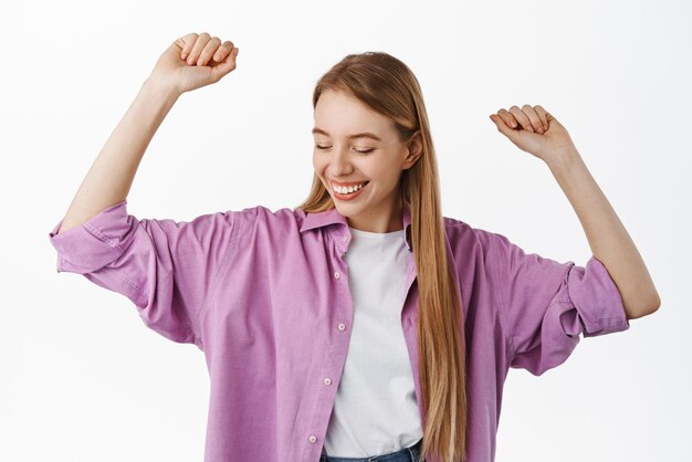 Fröhliches blondes Mädchen, das sorglos die Hände hochhebt und glücklich lächelt, während es Musik auf der Tanzfläche genießt, die vor weißem Hintergrund posiert