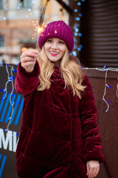 Fröhliches blondes Mädchen in stilvoller Kleidung, das leuchtende Wunderkerzen auf dem Weihnachtsmarkt in Kiew hält