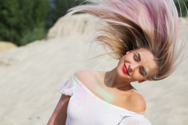 Fröhliches blondes Mädchen mit flatternden Haaren bedeckt mit trockener Farbe Holi in der Wüste. Platz für Text