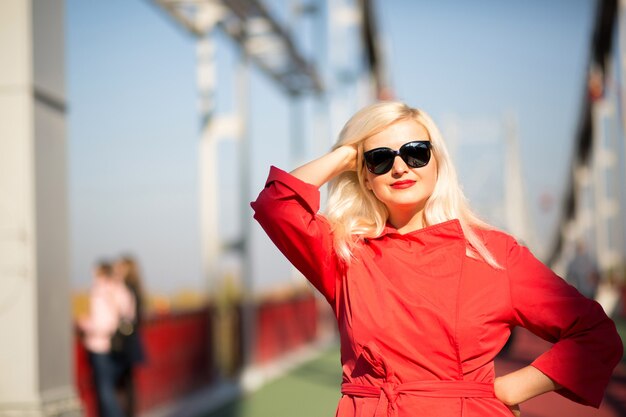 Fröhliches blondes Model posiert in rotem Mantel, trägt eine Sonnenbrille auf einem verschwommenen Stadthintergrund