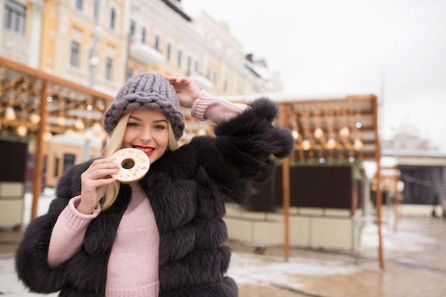 Fröhliches blondes Modell, das köstliche Ingwerplätzchen gegen Lichtdekoration auf dem Weihnachtsmarkt in Kiew hält