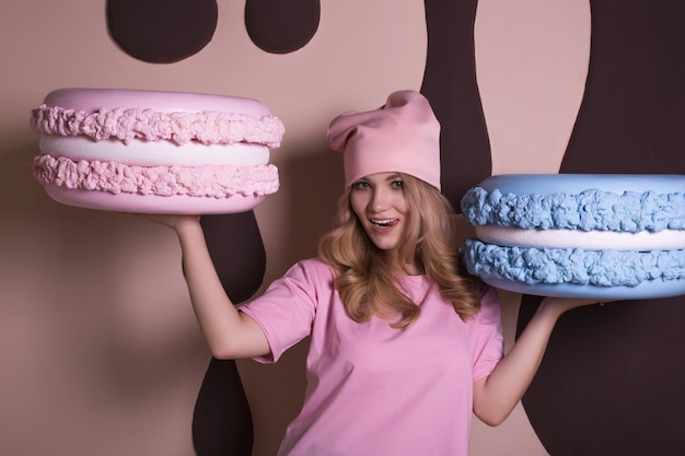 Fröhliches blondes Modell in rosa T-Shirt und Mütze mit großen leckeren Kuchenmakronen