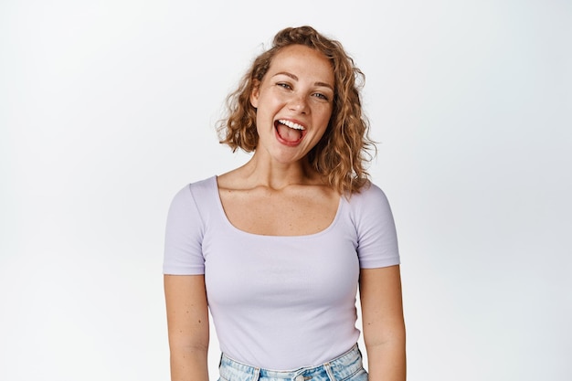 Fröhliches blondes weibliches Modell, das lacht und lächelt, glücklich in die Kamera schaut und im T-Shirt auf weißem Hintergrund steht. Platz kopieren