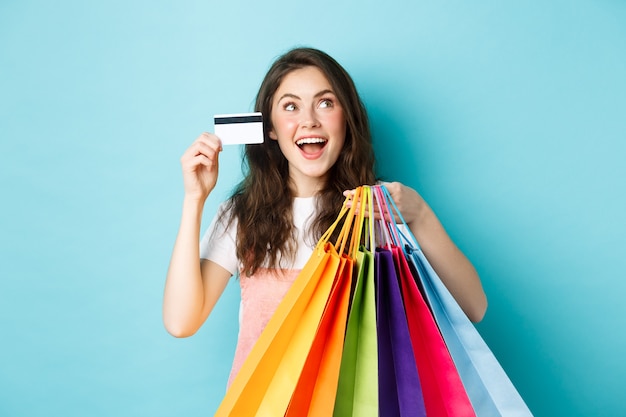 Fröhliches brünettes Mädchen, Shopaholic, das ihre Plastikkreditkarte und ihre Einkaufstaschen zeigt, schaut verträumt auf das Logo oder das Banner des Ladens, das auf blauem Hintergrund steht.