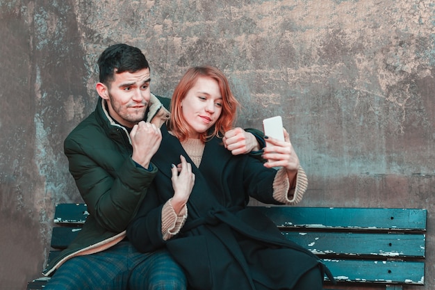 Fröhliches emotionales junges Paar, das auf der Bank sitzt und Selfie macht. Zwei glückliche Menschen Liebesgeschichte auf der Straße