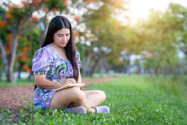 Fröhliches entspannendes Porträt einer freiberuflich tätigen Asiatin Tragen Sie während der Arbeit ein lila Kleid und halten Sie ein Tagebuch, während Sie im Freien auf einem grünen Rasen neben einem Stausee im Stadtpark sitzen