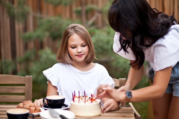 Fröhliches entzückendes Mädchen mit Mutter feiert mit Geburtstagskuchen auf der Café-Terrasse. 10 Jahre alt feiern Geburtstag.