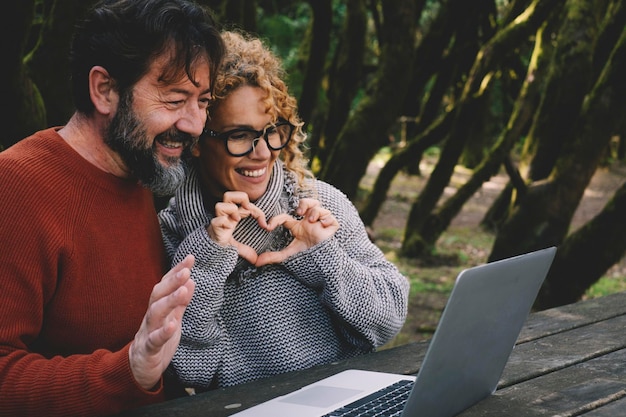 Fröhliches erwachsenes Paar hat Spaß bei der Videoanrufverbindung mit einem Laptop-Computer in der Freizeit im Freien in der Natur mit moderner Technologie Mann und Frau machen eine Herzgeste, um den Notebook-PC anzuzeigen