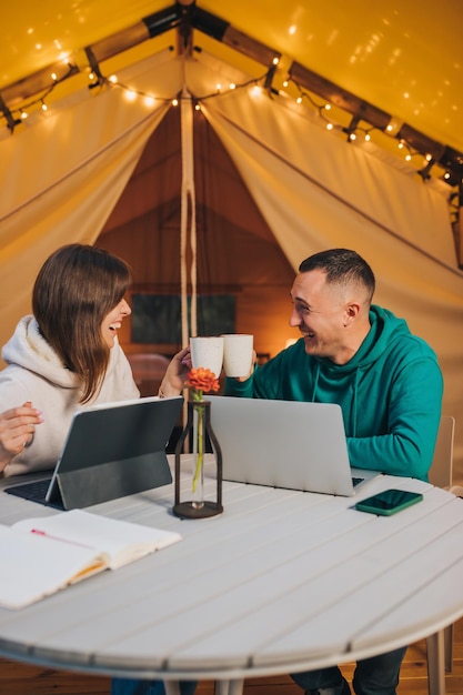 Fröhliches Familienpaar Freiberufler, die am Sommerabend in einem gemütlichen Glamping-Zelt mit Laptop arbeiten Luxus-Campingzelt für Urlaub im Freien und Urlaub Lifestyle-Konzept