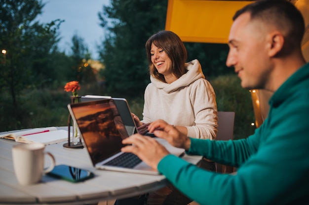Fröhliches Familienpaar, Freiberufler, die am Sommerabend in einem gemütlichen Glamping-Zelt mit Laptop arbeiten Luxus-Campingzelt für Urlaub im Freien und Urlaub Lifestyle-Konzept
