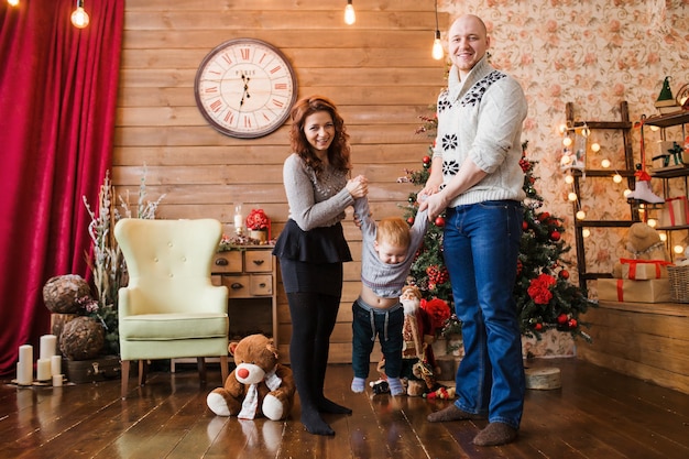 Fröhliches Familienporträt zu Weihnachten, Mutter, Vater und Sohn, die zu Hause auf einem Hochstuhl sitzen, Weihnachtsdekoration und Geschenke um sie herum