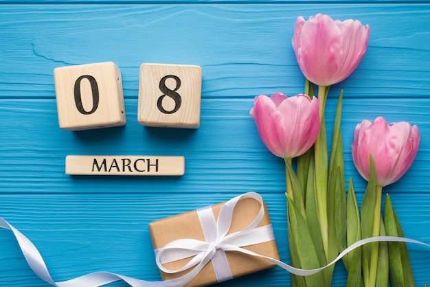 Fröhliches Fotokonzept zum internationalen Frauentag. Flatlay-Foto von zartrosa Tulpen verpackte Geschenkbox mit seidigem Band und Kalender isoliert heller Schreibtischtisch