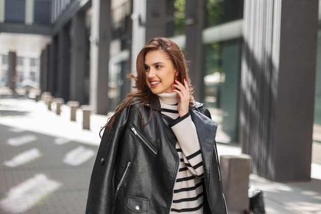 Fröhliches, fröhliches, schönes, stilvolles Frauenmodell in modischer Kleidung mit Lederjacke und gestreiftem Pullover geht an einem sonnigen Tag auf der Straße in der Nähe eines modernen Gebäudes spazieren