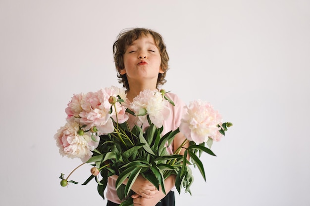 Fröhliches, glückliches Kind mit Pfingstrosen-Bouquet Lächelnder kleiner Junge auf weißem Hintergrund Muttertag Liebe und romantisches Konzept