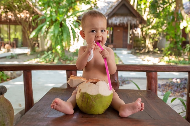 fröhliches glückliches kleines Baby sitzt auf dem Tisch und trinkt Kokosmilch aus frischer grüner Kokosnuss durch Stroh