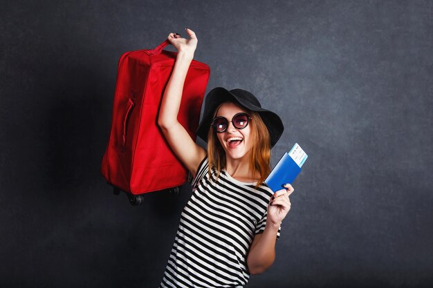 Fröhliches, glückliches Mädchen trägt einen schwarzen Hut und eine Sonnenbrille, die ein Passflugticket und einen roten Koffer vor grauem Hintergrund hält