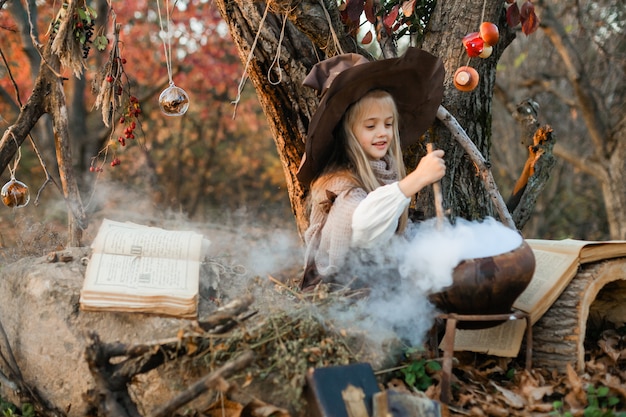 Fröhliches Halloween. Ein süßes Mädchen in einem Hexenkostüm ist in der Hexenhöhle. Nette fröhliche kleine Hexe braut einen Zaubertrank. Halloween.