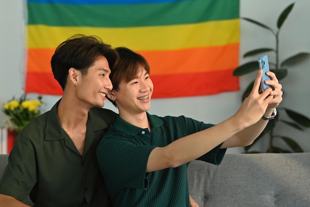 Fröhliches homosexuelles Paar, das im Wohnzimmer ein Selfie mit dem Smartphone macht, mit Regenbogenfahne im Hintergrund LGBT- und Liebeskonzept